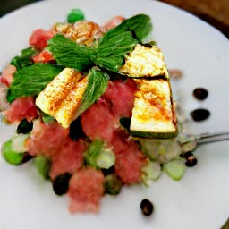 a salad with olive oil, spearmint, lemon zest, lemon juice, toasted seeds, and diced cucumbers. the salad is topped with fried halloumi cheese, watermelon, and basil