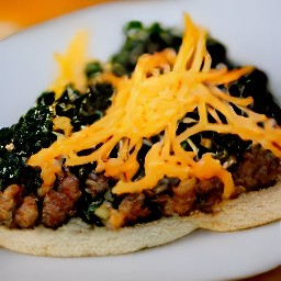 there are vegan refried beans, tomatoes, cooked crumbled meatless burgers patties, coated kale, and shredded cheddar cheese on corn tortillas on a plate.